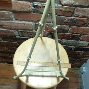 May include: A light green metal easel stand with a ruler on top. The easel is on a wooden stool.