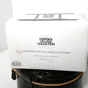 Puede incluir: Una caja blanca con el texto "HERITAGE VILLAGE COLLECTION" y "VICTORIA STATION TRAIN PLATFORM". La caja es un accesorio de porcelana pintado a mano. La caja está sobre un estuche de cuero negro.