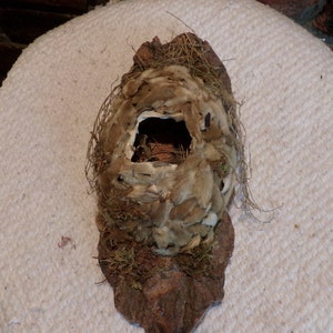 May include: A bird's nest made of twigs, moss, and feathers with a small opening in the centre. The nest is brown and white and is sitting on a white surface.