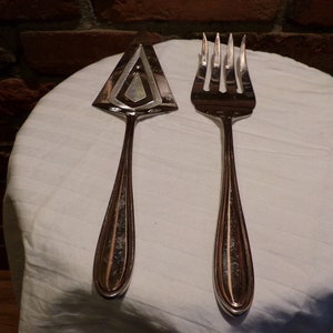 Puede incluir: Un juego de cuchillo para pastel y tenedor de plata. El cuchillo tiene una hoja triangular con un diseño decorativo. El tenedor tiene un diseño tradicional con cinco dientes.