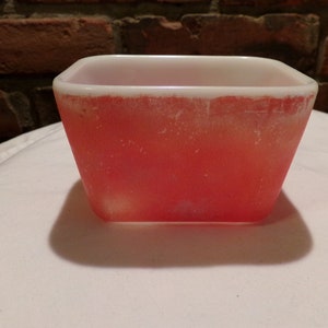 Puede incluir: Un cuenco cuadrado de vidrio rojo y blanco vintage. El cuenco tiene un borde blanco y un cuerpo rojo. El cuenco está sobre una superficie blanca, con una pared de ladrillo al fondo.