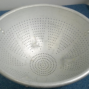 May include: A silver metal colander with many small holes for draining liquids. It features two curved handles. The round colander is shown on a blue surface. The diameter is approximately 25 cm.