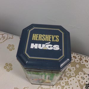 May include: A blue and gold tin with the text "HERSHEY'S HUGS" on the lid. The tin has a picture of a green and white scene on the front.