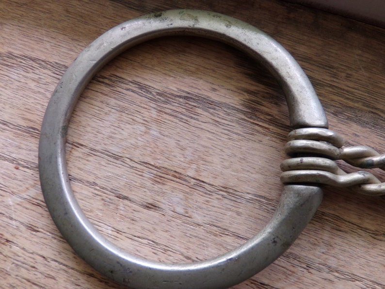 May include: A silver metal ring with a braided chain attached. The ring is smooth and has a rounded shape.