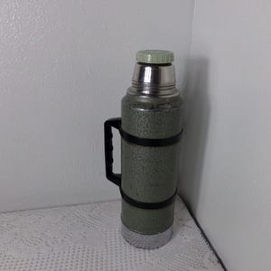 May include: A green and silver thermos with a black handle. The thermos has a textured surface and a silver lid.