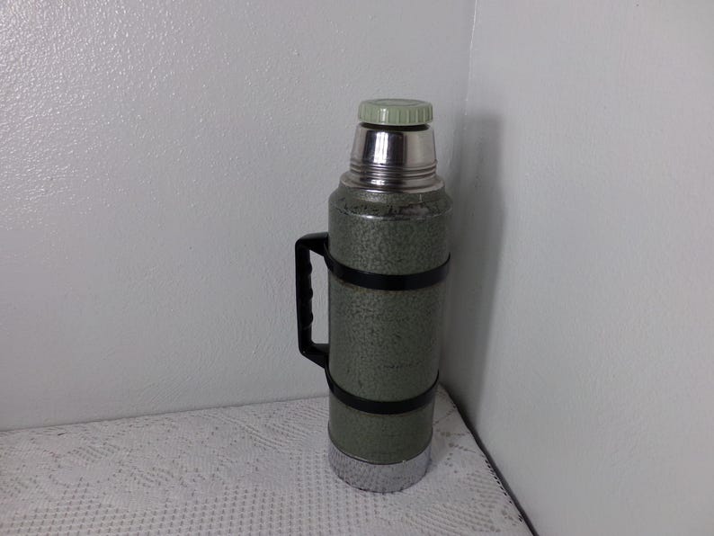 May include: A green and silver thermos with a black handle. The thermos has a textured surface and a silver lid.
