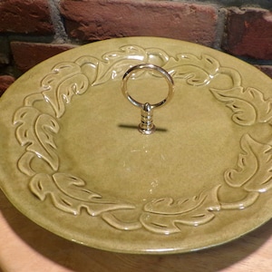 Op de afbeelding: Een groene keramische serveerschaal met een gouden handvat. De schaal heeft een reliëfdesign van bladeren rond de rand.