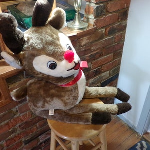 Può includere: Un grande renna di peluche marrone con un fiocco rosso e un naso rosso seduto su uno sgabello di legno. La renna ha grandi occhi marroni e un viso bianco.