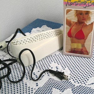 Pode incluir: Um dispositivo de cinto vibratório branco vintage com uma superfície texturizada e um cabo de alimentação preto. Uma caixa com as palavras "Vibrating Belt" e uma imagem de uma mulher de biquíni vermelho está ao lado do dispositivo.