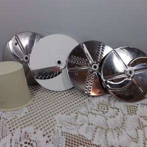 May include: Four different food processor blades, including a white plastic blade, a silver blade with a grating surface, and two silver blades with different slicing surfaces. The blades are on a white lace tablecloth.