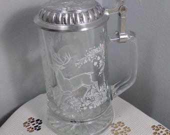 May include: A clear glass beer stein with a silver lid and handle. The stein features an etched design of a deer in a forest setting.