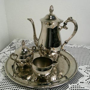 May include: A polished silver-tone tea service set consisting of a tall teapot with a lid, a lidded sugar bowl, and an open creamer pitcher. All three pieces are arranged on a round, matching silver-tone serving tray with a decorative beaded edge.