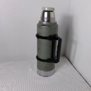 May include: A green metal thermos with a black handle and a silver lid. The thermos has a textured surface and a silver band around the middle.