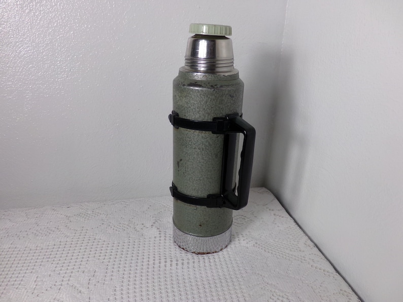 May include: A green metal thermos with a black handle and a silver lid. The thermos has a textured surface and a silver band around the middle.