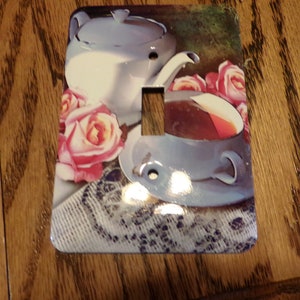 May include: A white porcelain teapot with a lid and a handle, a blue and white teacup with a saucer, and pink roses on a white lace tablecloth. The image is on a light switch cover.