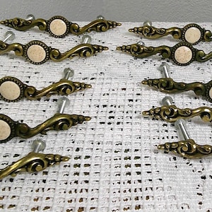 May include: A collection of antique brass drawer pulls. Each pull features an ornate design with a cream-colored center and a silver mounting post. The pulls are arranged on a white lace tablecloth.