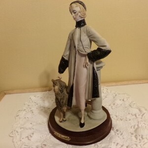 Pu&ograve; includere: Una statuetta in porcellana di una donna con un lungo cappotto con bordatura in pelliccia nera, in piedi con un piccolo cane. La statuetta &egrave; su una base di legno con il nome "Lladro" inciso su di essa.