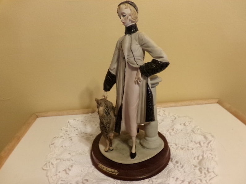 Pu&ograve; includere: Una statuetta in porcellana di una donna con un lungo cappotto con bordatura in pelliccia nera, in piedi con un piccolo cane. La statuetta &egrave; su una base di legno con il nome "Lladro" inciso su di essa.