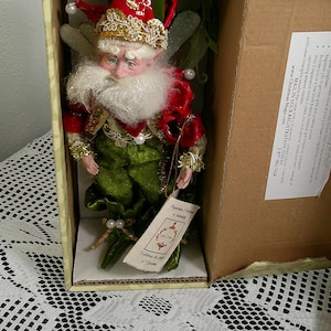 May include: A Christmas elf doll in a cardboard box. The elf is dressed in a red jacket, green pants, and a gold-trimmed hat. It has a white beard and is holding a tag. The box is open, revealing the doll inside.