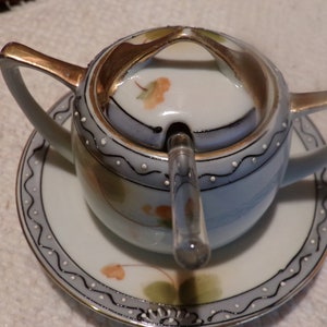 Op de afbeelding: Een witte porseleinen suikerpot met een deksel en een glazen lepel. De pot heeft een gouden rand en een bloemenmotief. Het deksel heeft een gouden handvat en een klein gaatje bovenaan.
