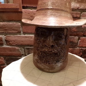 Puede incluir: Una escultura de cerámica de un rostro humano con un sombrero de ala ancha. La escultura es de color marrón y tiene una superficie texturizada. La escultura está sentada sobre una mesa blanca con un patrón de cuadrícula.