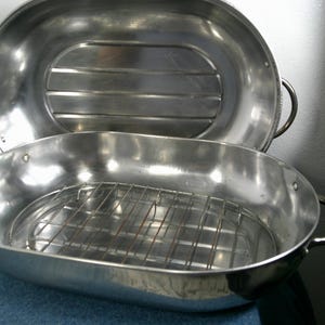 May include: An oval stainless steel roasting pan with a matching lid and a removable wire rack insert. The pan features two side handles and a polished metallic finish, with the lid showing horizontal ridges on the interior surface.