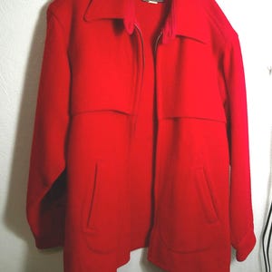 May include: A bright red jacket featuring a full-length front zipper, a wide collar, and two vertical slit pockets on the lower front. The garment has a horizontal seam across the chest and long sleeves, with a dark label visible at the neckline.