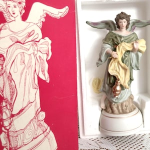 May include: A ceramic angel figurine in a white box with a red box next to it. The angel wears a green and gold robe and holds a yellow cloth and a lantern. The angel has brown hair and green wings. The box has a drawing of an angel.