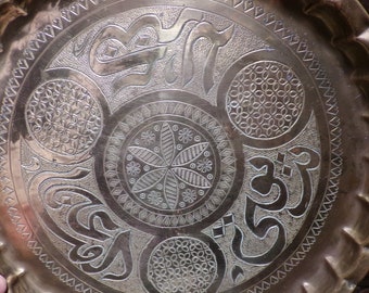 Puede incluir: Una bandeja redonda de servir de latón con un diseño intrincado grabado. El diseño presenta un motivo floral central rodeado de cuatro patrones geométricos más pequeños. La bandeja tiene un borde festoneado.
