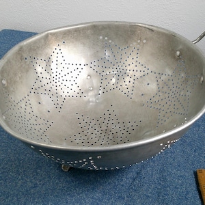 May include: A vintage, silver-colored metal colander with two handles. The colander has a bowl shape with numerous small holes and star-shaped patterns. It is sitting on a blue surface.