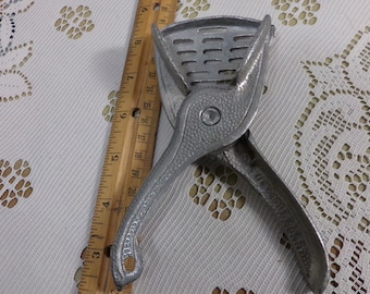 May include: A vintage, silver-colored citrus juicer with a hinged design. The juicer features a slotted pressing surface and long handles for leverage. A wooden ruler is placed next to the juicer, providing a size reference.