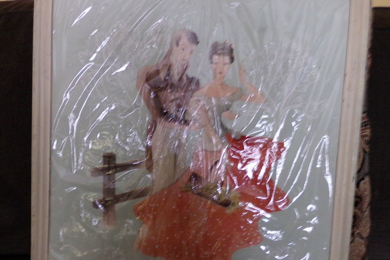 May include: A framed image of a couple standing together. The man is wearing a brown shirt and the woman is wearing a red dress. The image is covered in plastic wrap.