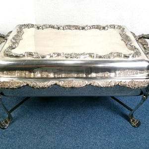 May include: A rectangular silver-plated chafing dish featuring an ornate, embossed floral border along the lid and base. The serving vessel is supported by four curved legs with decorative feet and includes two side handles for easy transport.