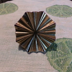 May include: A silver-toned, octagonal-shaped decorative knob with a fan-like design. The knob has a smooth, shiny finish.