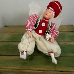 Könnte beinhalten: Eine Puppe mit einem rot-weiß gestreiften Langarmshirt, einer roten Weste mit zwei weißen Knöpfen und einer cremefarbenen, weiten Hose. Die Puppe trägt eine rote Mütze mit weißem Rand, hat langes weißes Haar und besitzt durchsichtige Glitzerflügel.