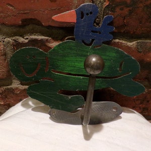 May include: A green and blue metal turtle hook with a bird on its back. The hook is attached to a wall with a brick background.