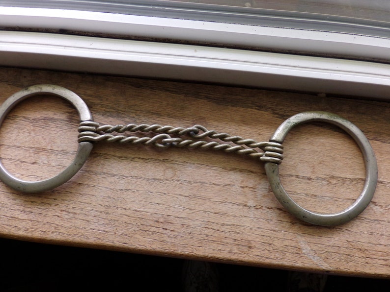 May include: A silver metal horse bit with a braided chain connecting two circular rings.