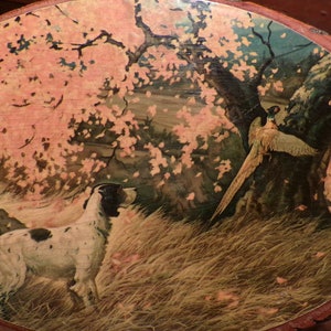 Op de afbeelding: Een ronde houten plaquette met een geschilderd tafereel van een witte en bruine hond die wijst naar een fazant in een bosrijke omgeving. De achtergrond toont een roze en groene boom met bruine takken.