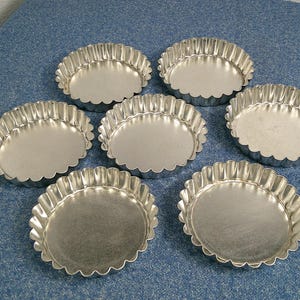 Puede incluir: Siete moldes pequeños y redondos para tartaletas, de metal plateado y con estrías. Los moldes están dispuestos sobre una superficie azul texturizada. Cada molde tiene un borde festoneado.