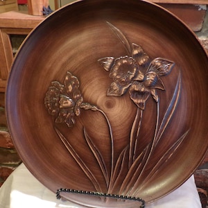 Puede incluir: Una bandeja redonda de madera con un diseño tallado de dos flores estilizadas con hojas largas. La bandeja es marrón y tiene un acabado brillante.