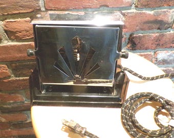 May include: A vintage black electric toaster with a decorative fan-shaped design on the front. The toaster has two knobs and a power cord with a plug. It sits on a light-colored surface against a brick wall.