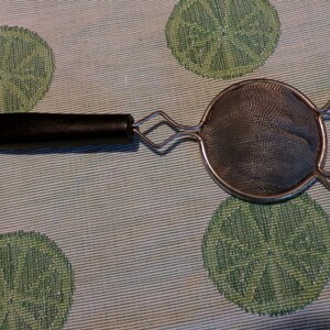 May include: A black handled, silver mesh tea strainer with a round shape and two curved handles.