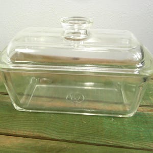 Pode incluir: Um recipiente retangular de vidro transparente para frigorífico com tampa a condizer. A tampa possui uma pega central em forma de botão. A base do recipiente tem um logótipo circular em relevo com um W no interior.