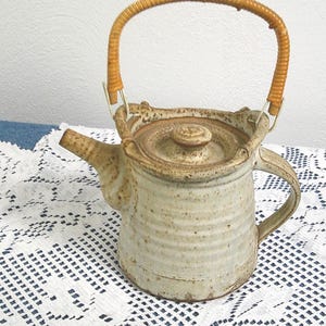 May include: A ceramic teapot with a light beige speckled finish. It features a rounded body, a spout, and a handle made of woven light brown material. The lid has a small knob. The teapot is sitting on a white lace tablecloth.