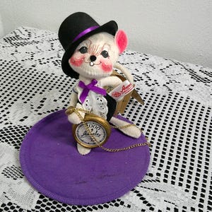 Op de afbeelding: Een Annalee-pop van een muis met een zwarte hoge hoed met een paarse band, een zwart vest en een wit overhemd met een paarse strik. De muis houdt een gouden zakhorloge vast en zit op een paarse vilten cirkel.