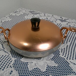 Op de afbeelding: Een koperkleurige metalen braadpan met een zilveren basis. De pan heeft twee koperen handvatten en een zwarte knop op het deksel. De pan staat op een wit kanten tafelkleed.