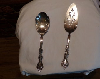 May include: Two silver serving utensils, a spoon and a cake server, with ornate handles. Both utensils have a decorative design.