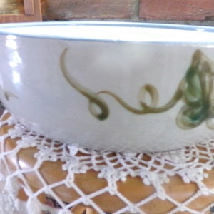 May include: A white ceramic bowl with a blue rim and green leaf design. The bowl is sitting on a crocheted doily.