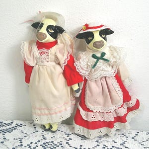 May include: Two cow-faced dolls wearing red and cream-colored dresses with white lace trim. One doll wears a hat with a pink ribbon, and the other wears a hat with a white ribbon. The dolls are standing side by side.