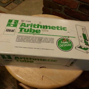 Może przedstawiać: Białe pudełko z zielonymi i czerwonymi akcentami dla edukacyjnej zabawki Ideal Arithmetic Tube. Pudełko zawiera zielono-białą ilustrację osoby wrzucającej zielone kulki do rury. Pudełko zawiera również tekst "No. 7725 Arithmetic Tube" i "Nowy projekt!"